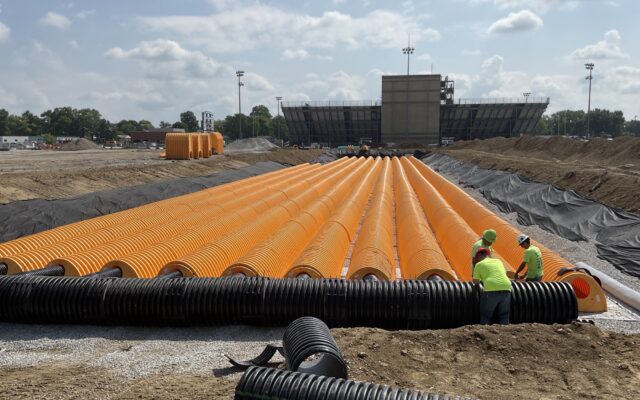 Workers install orange and black drainage pipes in trench near stadium.