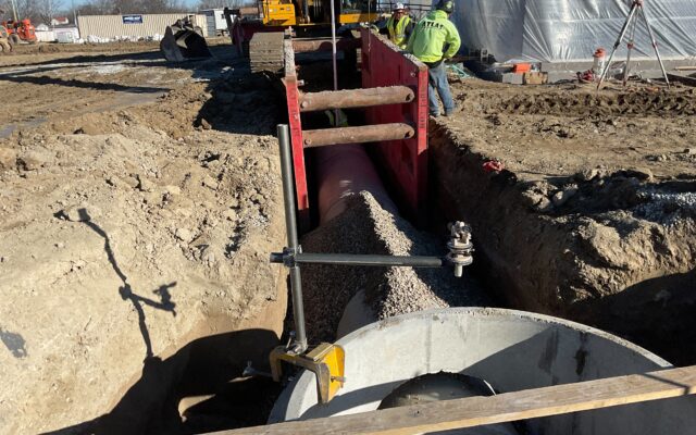 Workers install underground pipe with gravel and trench shoring.