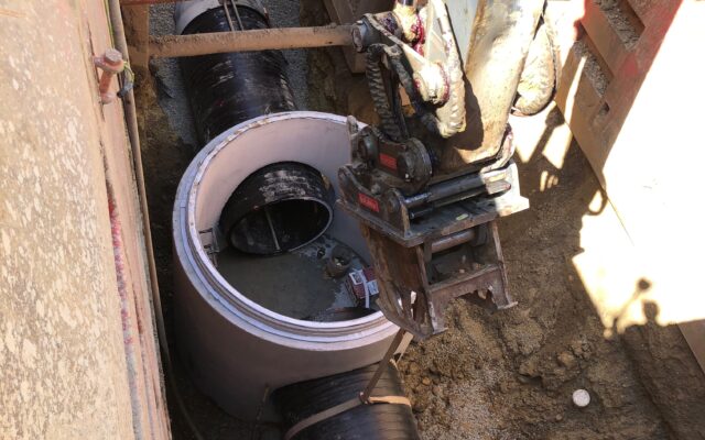Worker inside concrete pipe in trench; excavator and rebar above.