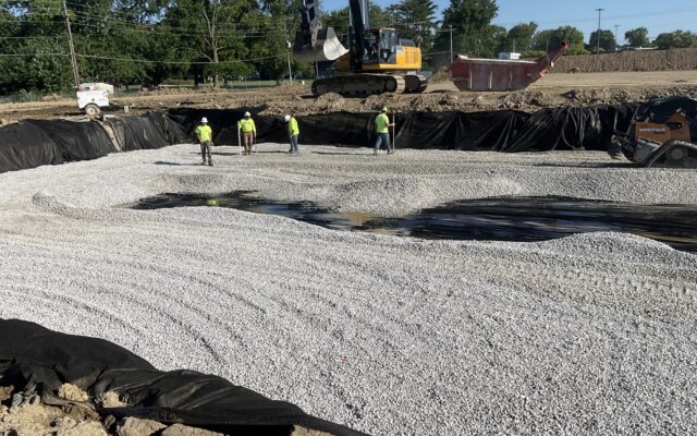 Four workers on gravel at a dig site with excavator.