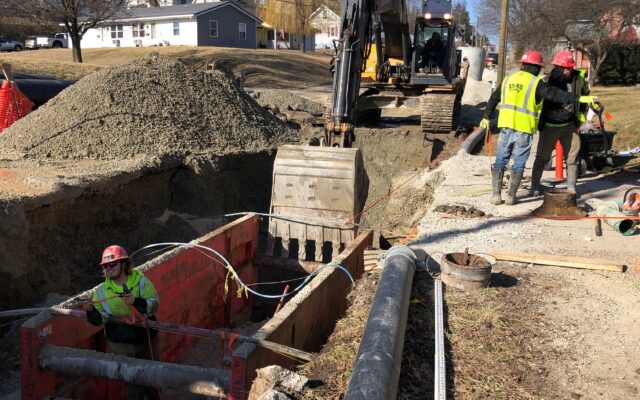 Workers install pipes with heavy machinery in a street trench.