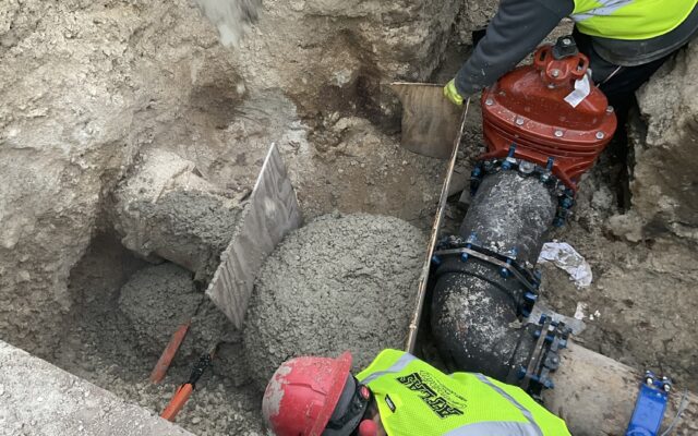 Two workers in safety gear secure pipes and pour concrete.