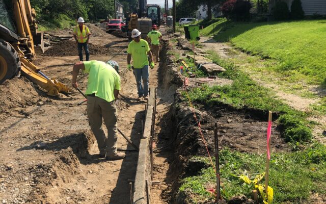 Workers dig trench on residential street with machinery nearby.