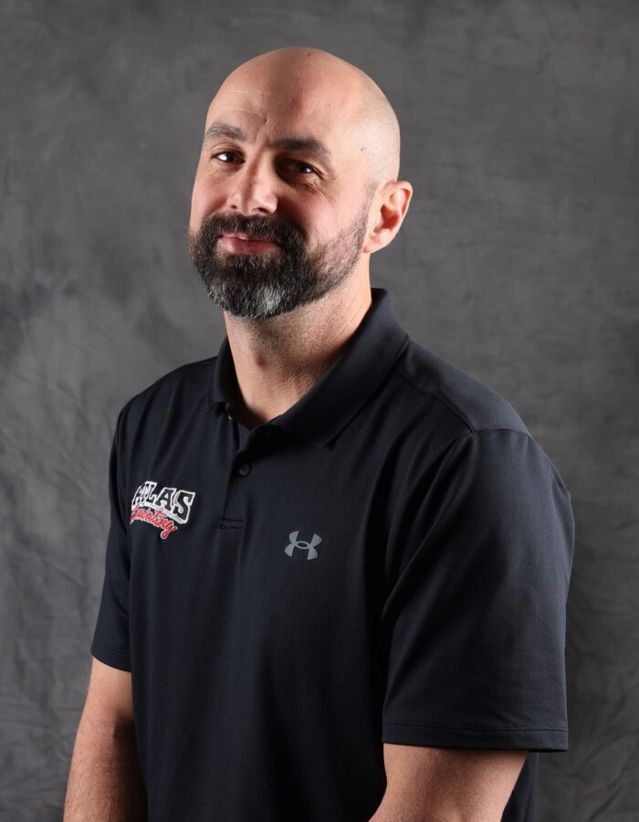 Bald bearded man in black polo before gray textured background.