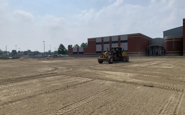 Grader on dirt site by modern brick school, clouds above.