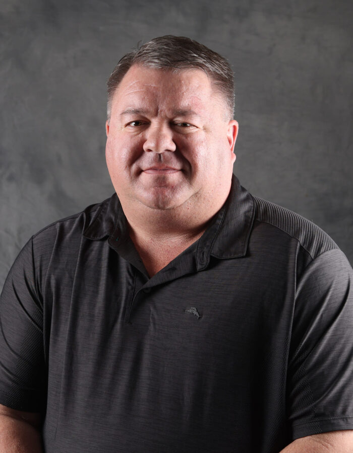 Man in dark polo shirt sits before a plain gray backdrop.