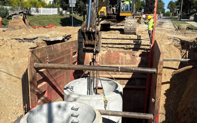 Excavator lowers concrete pipe into trench as workers assist.