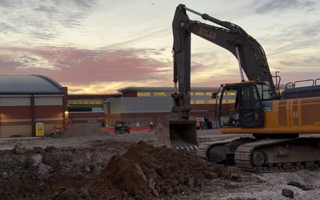 Excavator on dirt site at sunset with cloudy sky and materials.