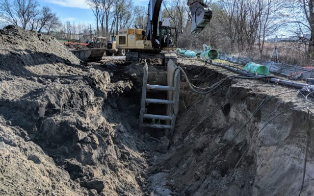 Excavator by deep trench with pipes and trench boxes at construction site.