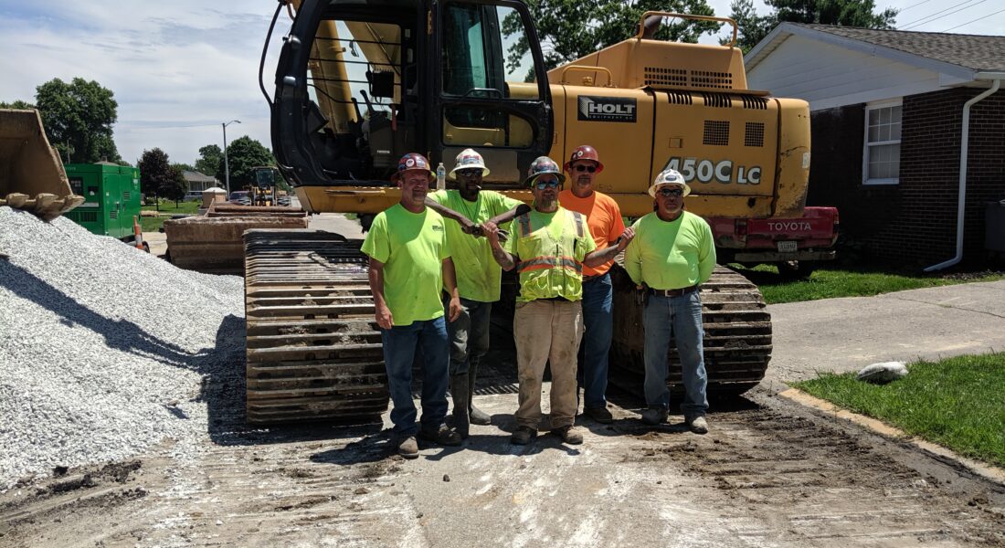 Five workers in safety gear stand by an excavator and gravel.