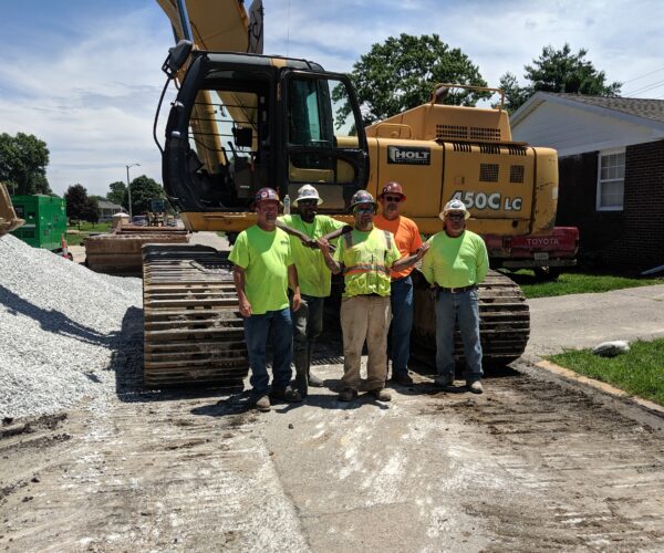 Five workers in safety gear stand by an excavator and gravel.