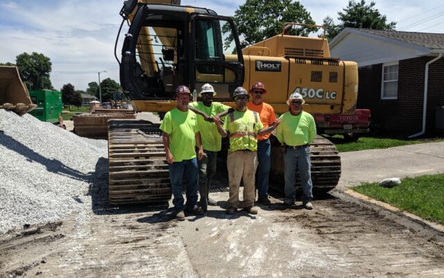 Five workers in safety gear stand by an excavator and gravel.