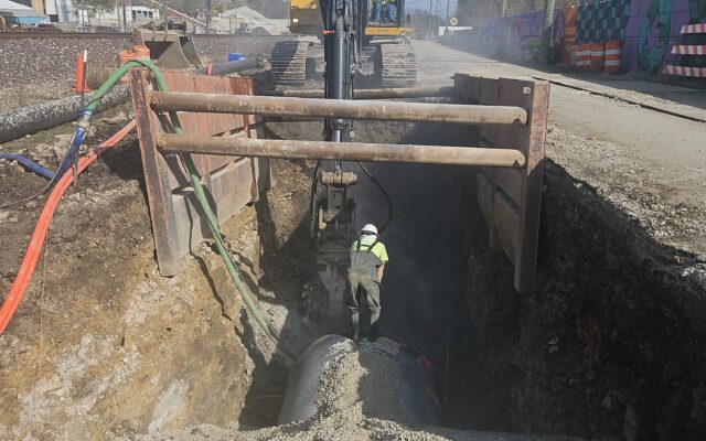 Worker in trench guides excavator placing equipment on large pipes.