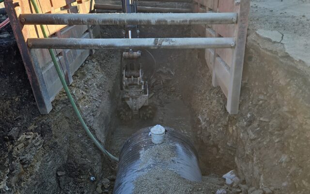 Excavator fills trench with gravel around large cylindrical tank.