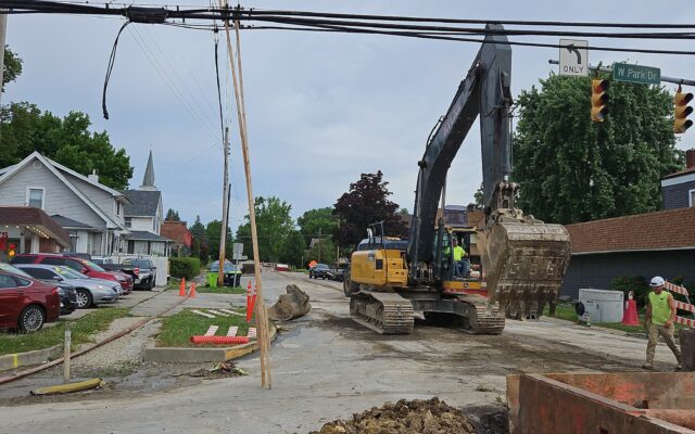 Crew using excavator on damaged street, houses and cars nearby.