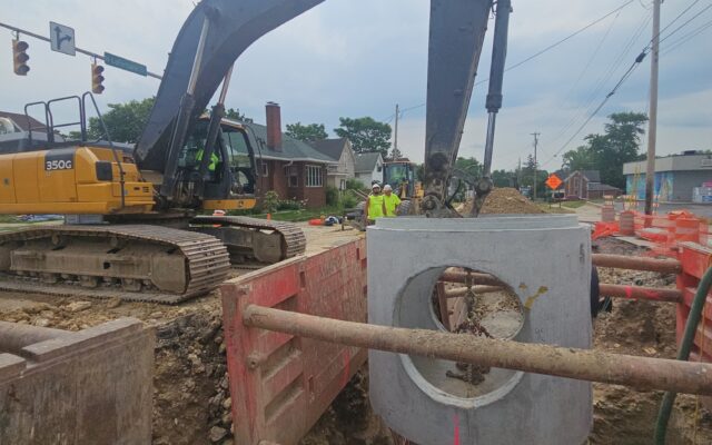 Workers watch excavator lifting pipe at construction site.