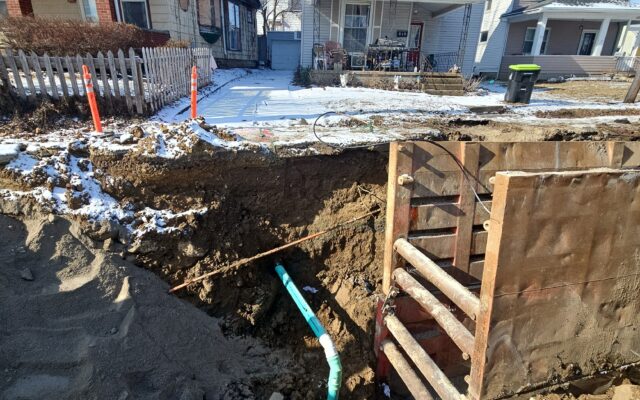 Hole in snowy yard exposes water pipes; barriers visible. Timestamped.