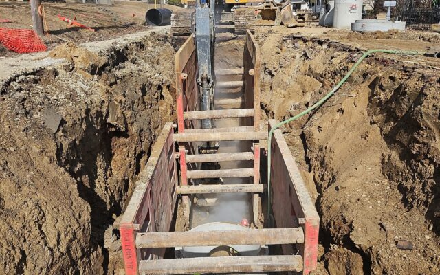 Excavator by deep shored trench on residential street; houses line both sides.