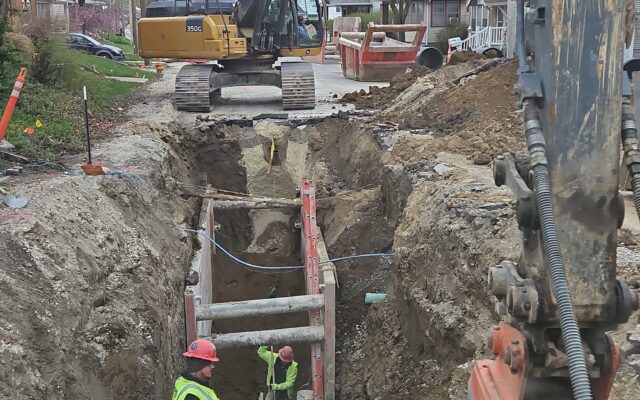 Workers in deep trench; excavator above on a residential street.