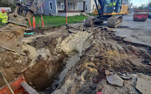 Excavator digging city street, pipes exposed, workers nearby.