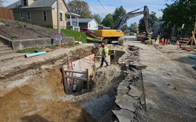 Worker by trench, excavator and barriers on street.