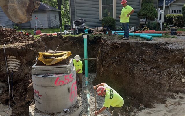 Workers install concrete sewer pipe as another observes; backhoe nearby.