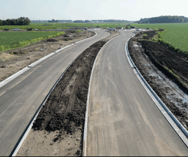Newly paved road under construction runs through green farmland.