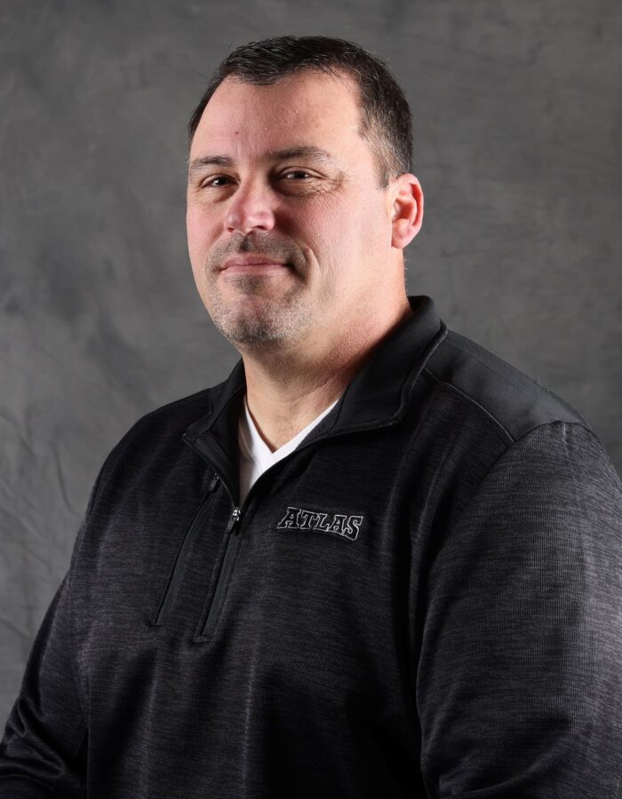 Man in black sweater and white tee before gray background.