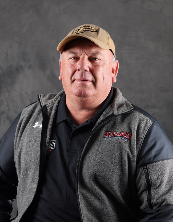 Man in ATLAS jacket and tan cap poses by gray backdrop.