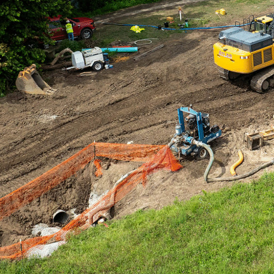 Excavator, orange fencing, trench, scattered equipment on dirt.
