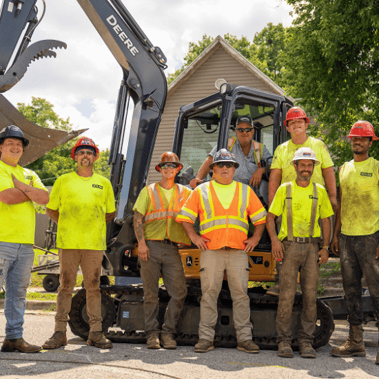 Seven construction workers in safety gear by a small excavator.