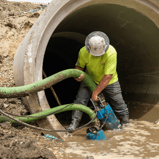 Worker connects hoses to blue pump by muddy concrete pipe.