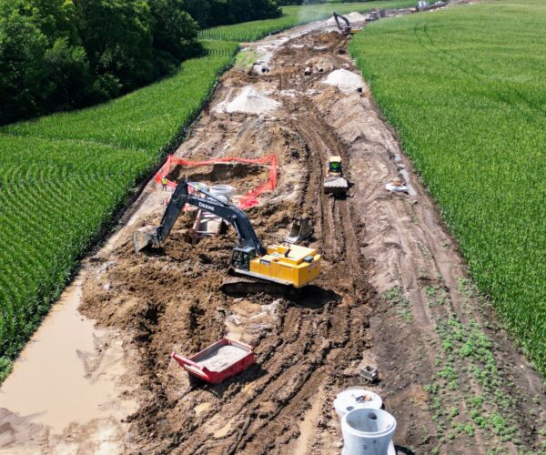 Excavators work on a muddy pipeline site between green fields.