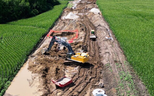 Excavators work on a muddy pipeline site between green fields.