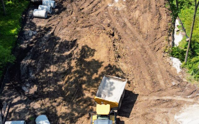 Aerial view of construction site with dump truck, excavator, trees.