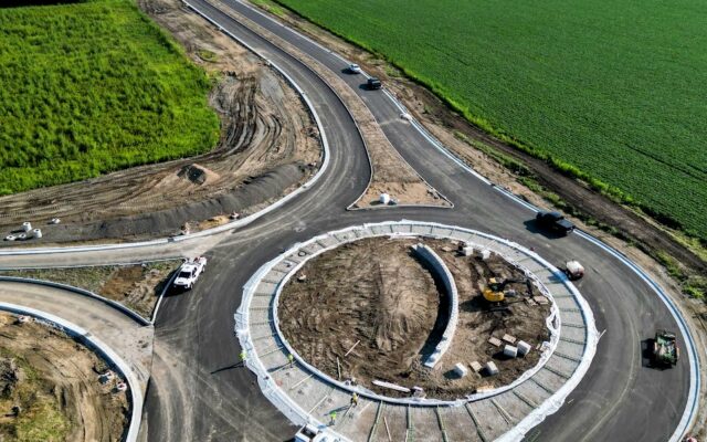 Roundabout under construction with vehicles, green fields around.