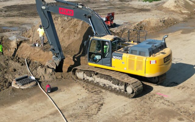 Yellow excavator digging at site with workers and houses behind.