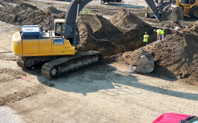 Yellow excavator digging with three workers; red vehicle in front.