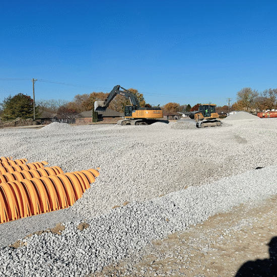 Construction site with gravel piles, two excavators, pipes, and trees.