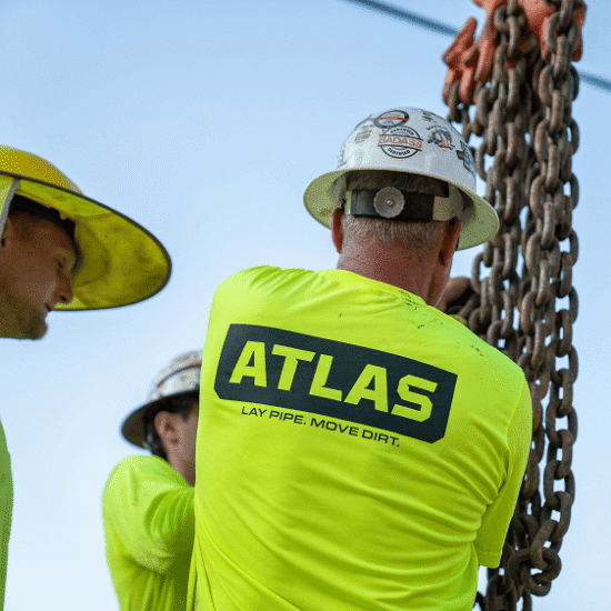 Three workers in helmets use chain; one wears ATLAS LAY PIPE MOVE DIRT shirt.