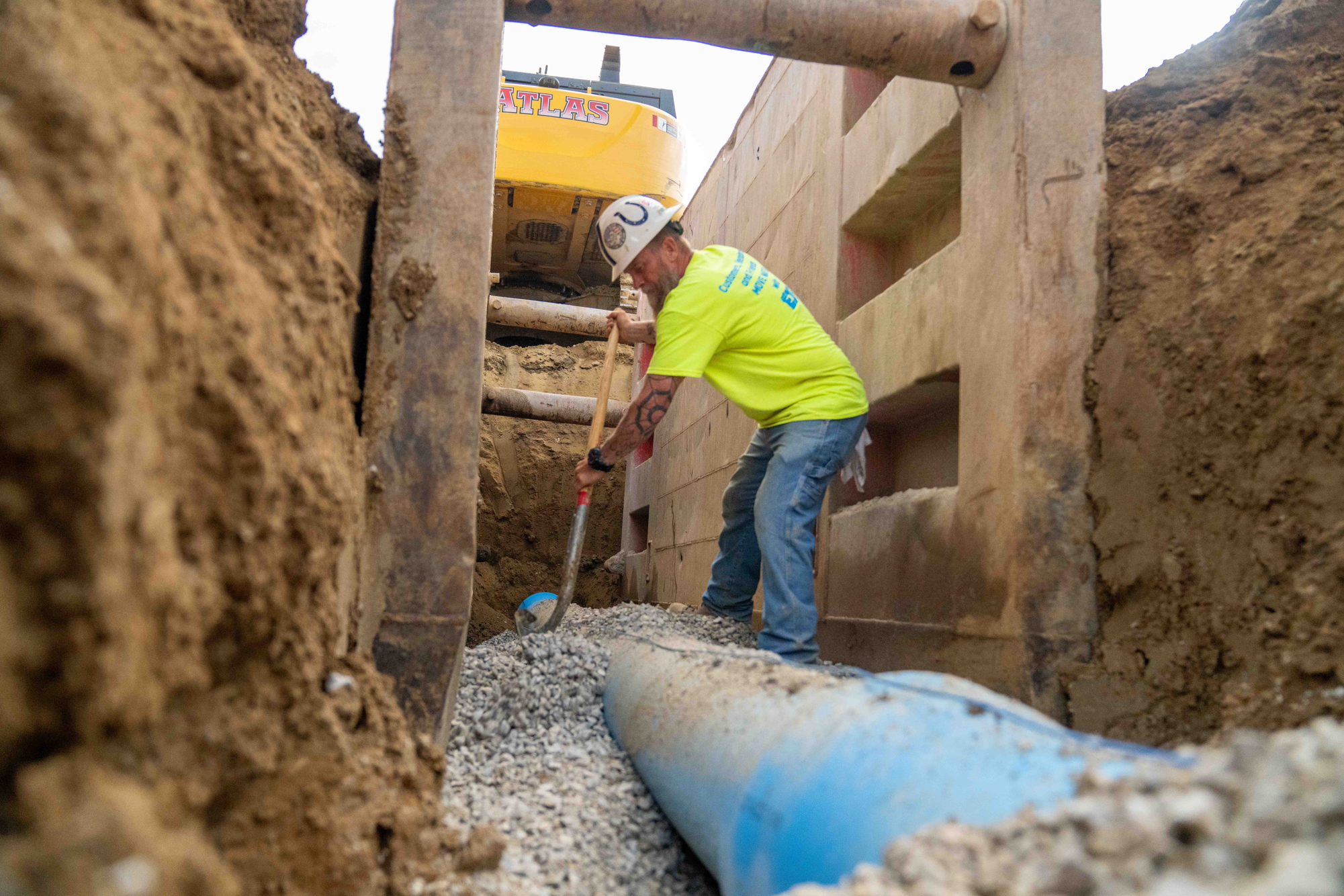 Worker in hard hat shovels gravel near large blue pipe.