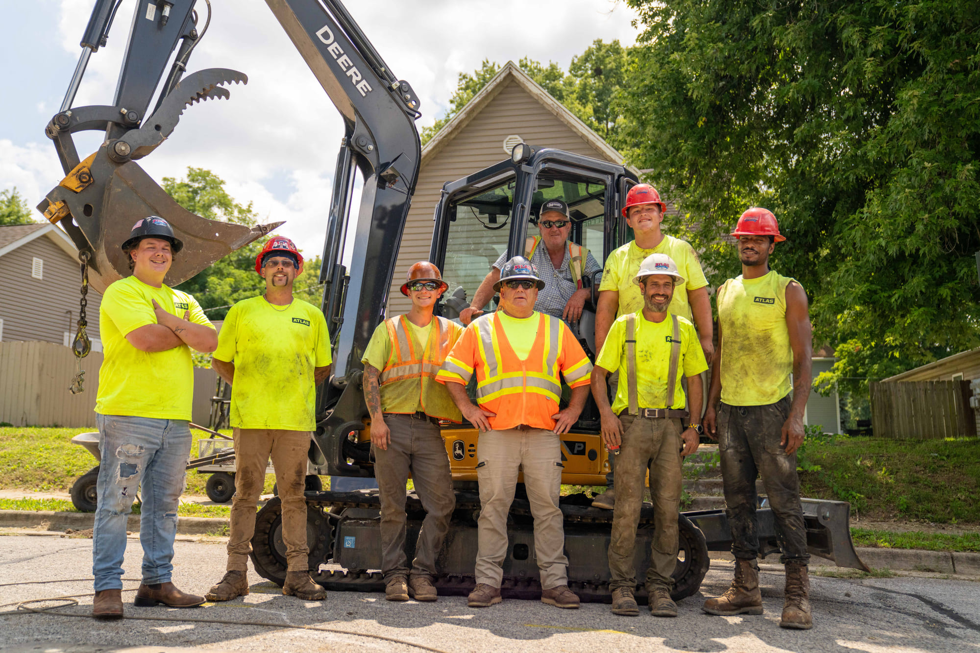 Eight construction workers in safety gear pose by a small excavator.
