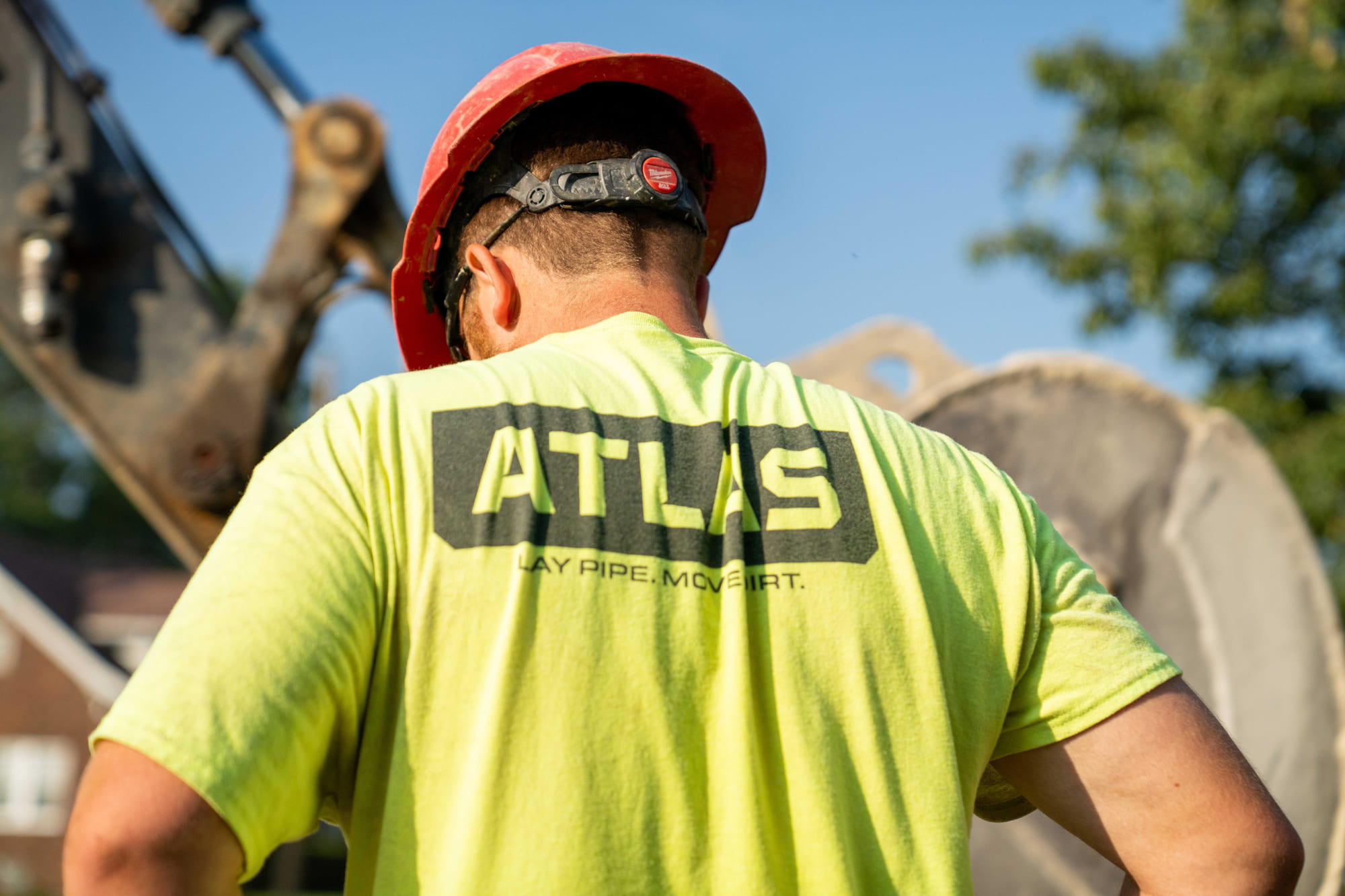 Construction worker in red helmet, yellow shirt by machines outside.