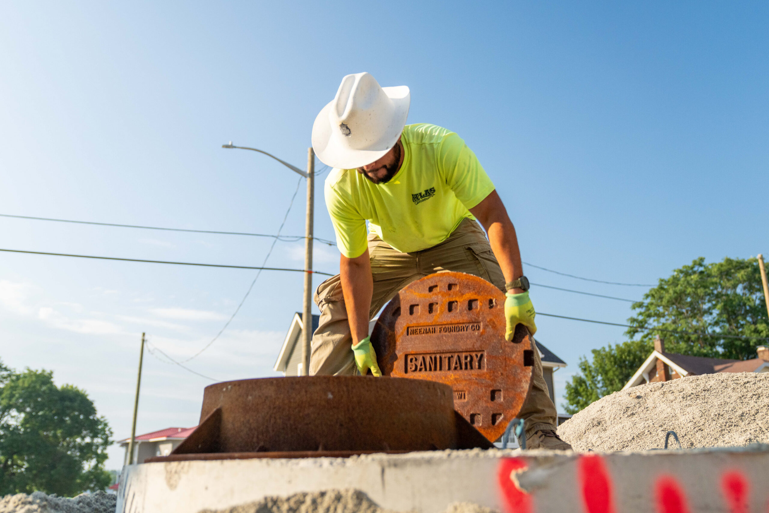 Worker in hard hat lifts manhole cover outdoors.