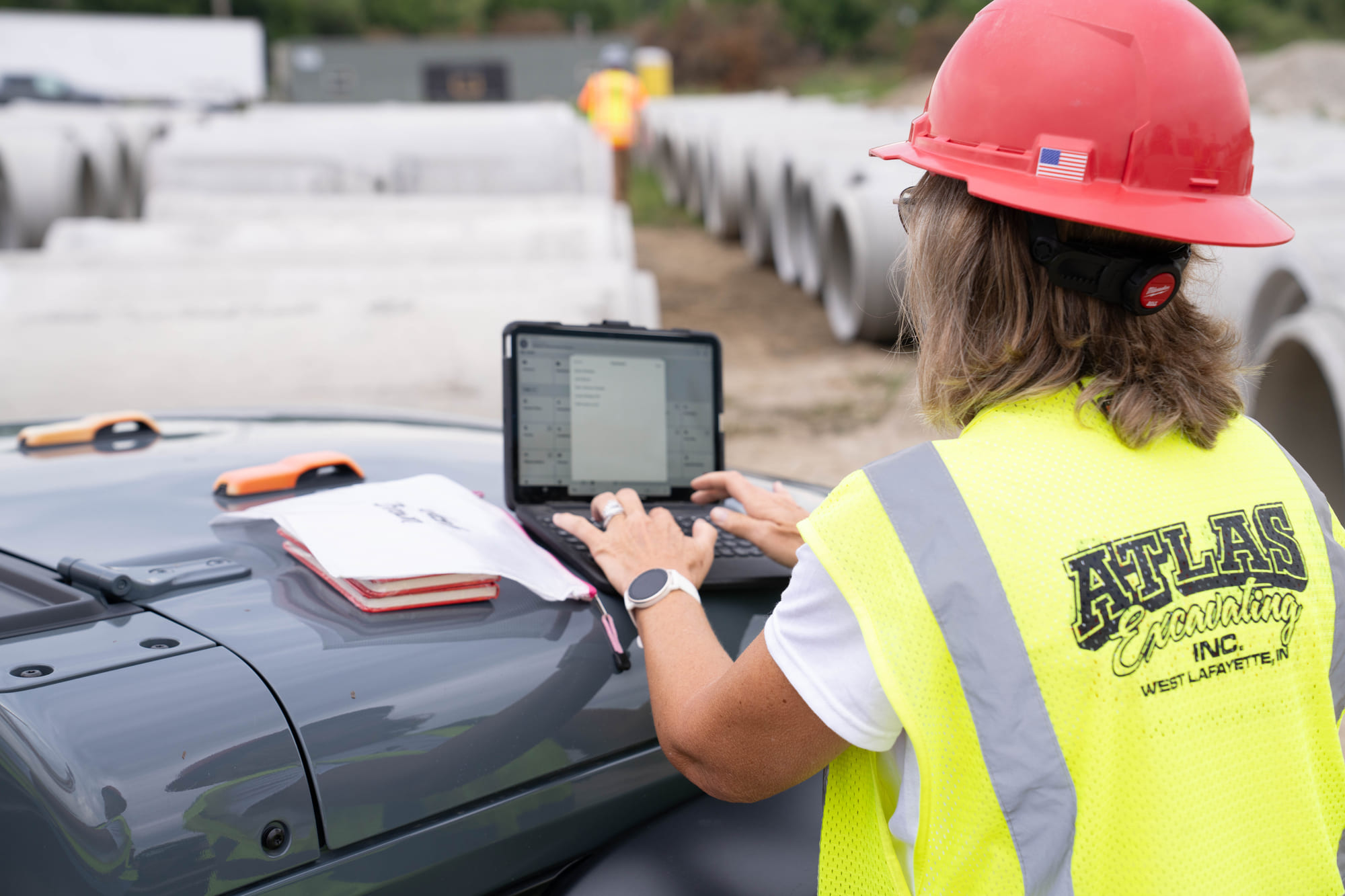 Worker in safety vest uses laptop on vehicle near concrete pipes.