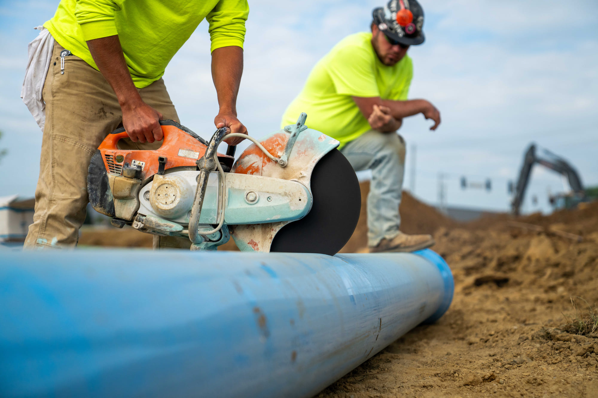 Worker cuts blue pipe with saw; another watches nearby.