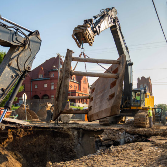Excavators lift steel trench box; historic red brick building behind.