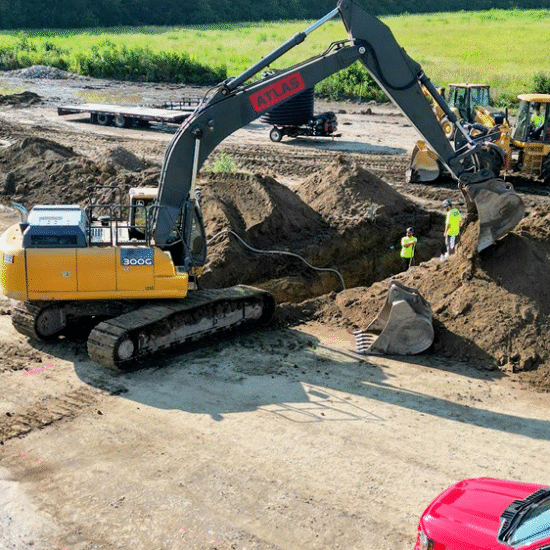 Excavator digs trench as two workers watch; bulldozer, trailer behind.