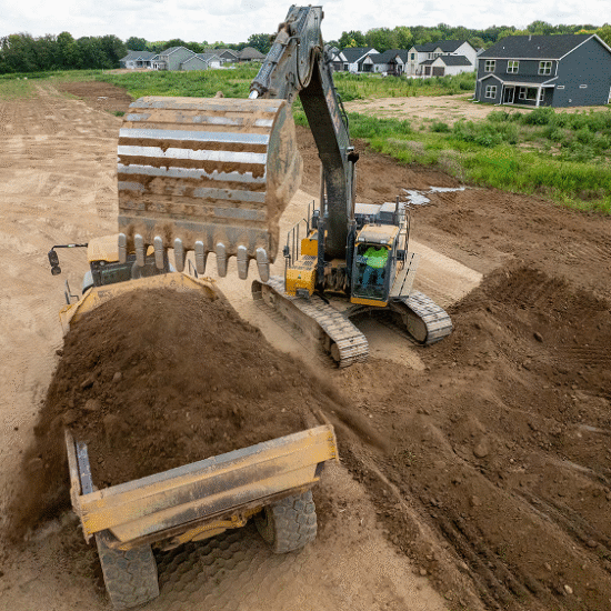 Excavator loading dirt into dump truck by houses.