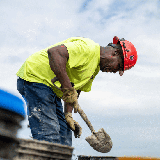 Worker in red hard hat shovels cement on a cloudy worksite.